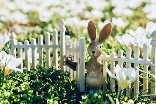 Cute bunny figurine beside a white fence in a lush spring garden with blooming white flowers.