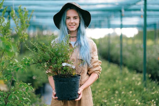 pexels-photo-5029807-5029807 A young woman smiles while holding a potted plant, standing in a greenhouse.